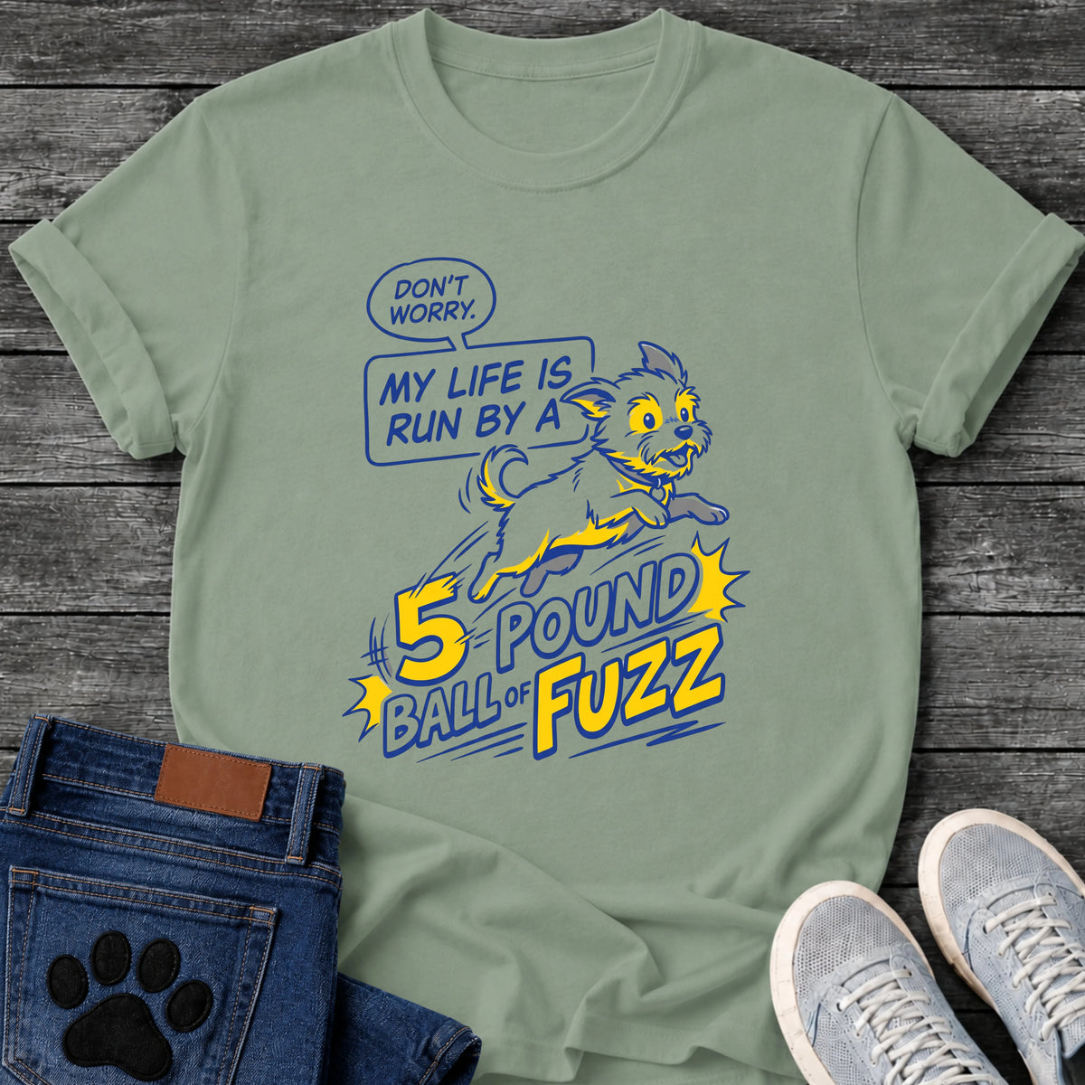 Illustrated graphic tee design of a small energetic dog leaping, with a speech bubble reading "Don't Worry, My Life Is Run by a 5 Pound Ball of Fuzz" in bold comic style type.
