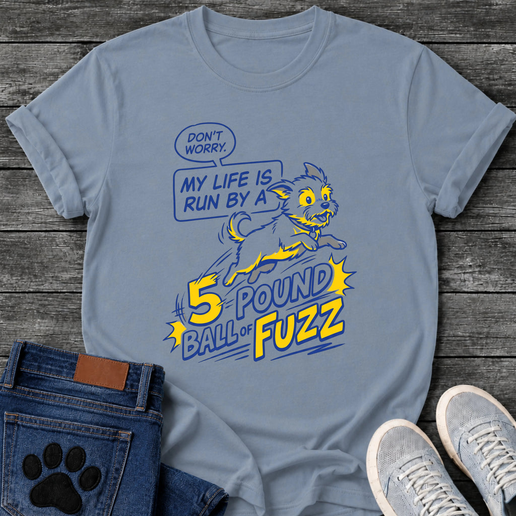 Illustrated graphic tee design of a small energetic dog leaping, with a speech bubble reading "Don't Worry, My Life Is Run by a 5 Pound Ball of Fuzz" in bold comic style type.