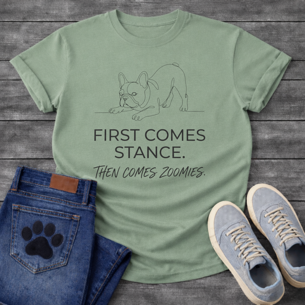 French Bulldog t-shirt with minimal continuous line art Frenchie in play bow zoomies stance, bold sans-serif text reading First Comes Stance Then Comes Zoomies, single dark ink on warm sand tee, premium clean design