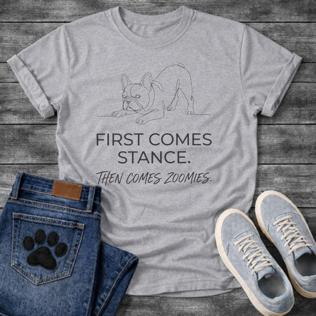 French Bulldog t-shirt with minimal continuous line art Frenchie in play bow zoomies stance, bold sans-serif text reading First Comes Stance Then Comes Zoomies, single dark ink on warm sand tee, premium clean design