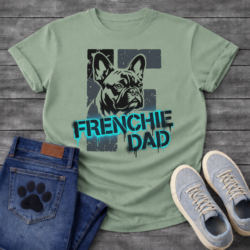 French Bulldog t-shirt with stencil-style Frenchie portrait over distressed dark block background, bold neon cyan graffiti text reading Frenchie Dad with black drip effects, high contrast urban street art graphic on warm sand tee

