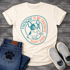 French Bulldog t-shirt with circular badge design, cartoon Frenchie holding a TV remote, teal and coral circular text reading Frenchie Is My Favorite Reality Show, two-color retro emblem on warm sand tee