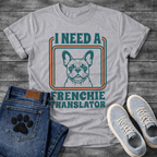 French Bulldog t-shirt with retro 70s groovy typography reading I Need a Frenchie Translator, detailed Frenchie portrait centered in teal and orange rounded rectangle frame, two-color retro illustration on warm sand tee
