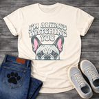 French Bulldog t-shirt with retro 70s bubble typography reading I'm Always Watching You above a peeking Frenchie face with pink inner ears, vintage halftone illustration on warm sand tee, groovy retro style