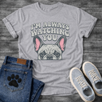 French Bulldog t-shirt with retro 70s bubble typography reading I'm Always Watching You above a peeking Frenchie face with pink inner ears, vintage halftone illustration on warm sand tee, groovy retro style