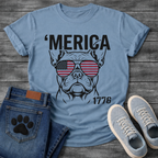 French Bulldog t-shirt with bold 'Merica text above a detailed Frenchie face wearing American flag aviator sunglasses, 1776 text bottom right, black and red patriotic graphic on warm sand tee