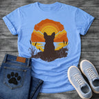 French Bulldog t-shirt with Frenchie silhouette seen from behind sitting on cracked earth beside water, cattail reeds on either side, bold concentric sun rings in deep orange and gold behind, distressed vintage texture throughout, organic blob frame, no text
