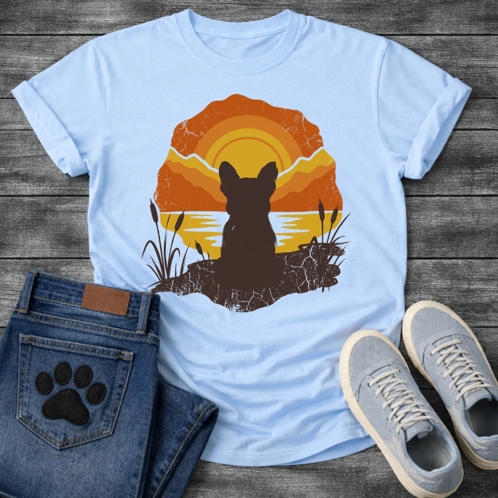 French Bulldog t-shirt with Frenchie silhouette seen from behind sitting on cracked earth beside water, cattail reeds on either side, bold concentric sun rings in deep orange and gold behind, distressed vintage texture throughout, organic blob frame, no text
