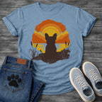  French Bulldog t-shirt with Frenchie silhouette seen from behind sitting on cracked earth beside water, cattail reeds on either side, bold concentric sun rings in deep orange and gold behind, distressed vintage texture throughout, organic blob frame, no text
