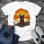French Bulldog t-shirt with Frenchie silhouette seen from behind sitting on cracked earth beside water, cattail reeds on either side, bold concentric sun rings in deep orange and gold behind, distressed vintage texture throughout, organic blob frame, no text
