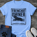 French Bulldog t-shirt with vintage distressed poster frame, bold oversized text reading Frenchie Owner above a splooting Frenchie lying flat on tile floor, bold banner text below reading Permanently Indoors, single dark ink halftone graphic with heavy crackle texture