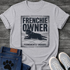 French Bulldog t-shirt with vintage distressed poster frame, bold oversized text reading Frenchie Owner above a splooting Frenchie lying flat on tile floor, bold banner text below reading Permanently Indoors, single dark ink halftone graphic with heavy crackle texture