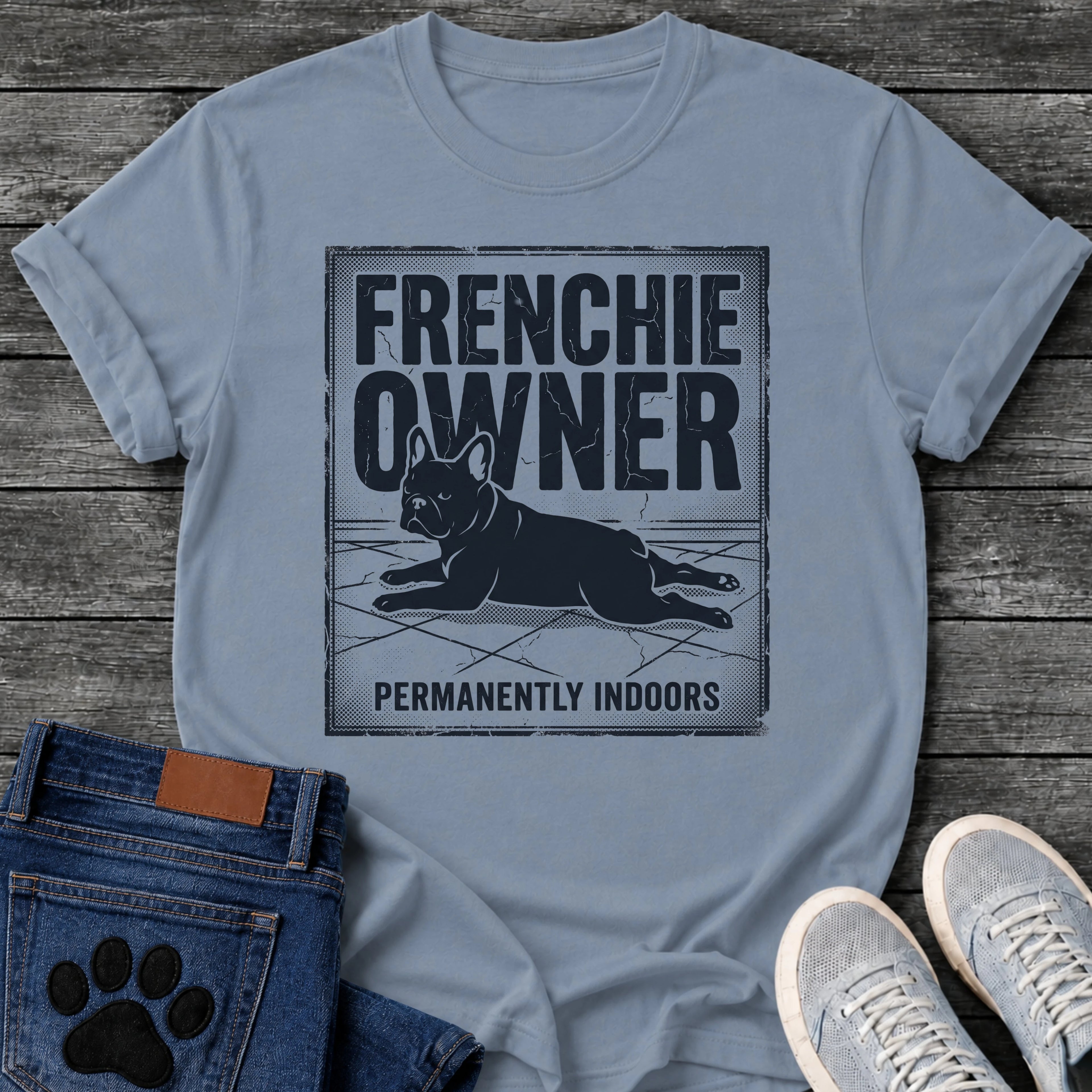 French Bulldog t-shirt with vintage distressed poster frame, bold oversized text reading Frenchie Owner above a splooting Frenchie lying flat on tile floor, bold banner text below reading Permanently Indoors, single dark ink halftone graphic with heavy crackle texture
