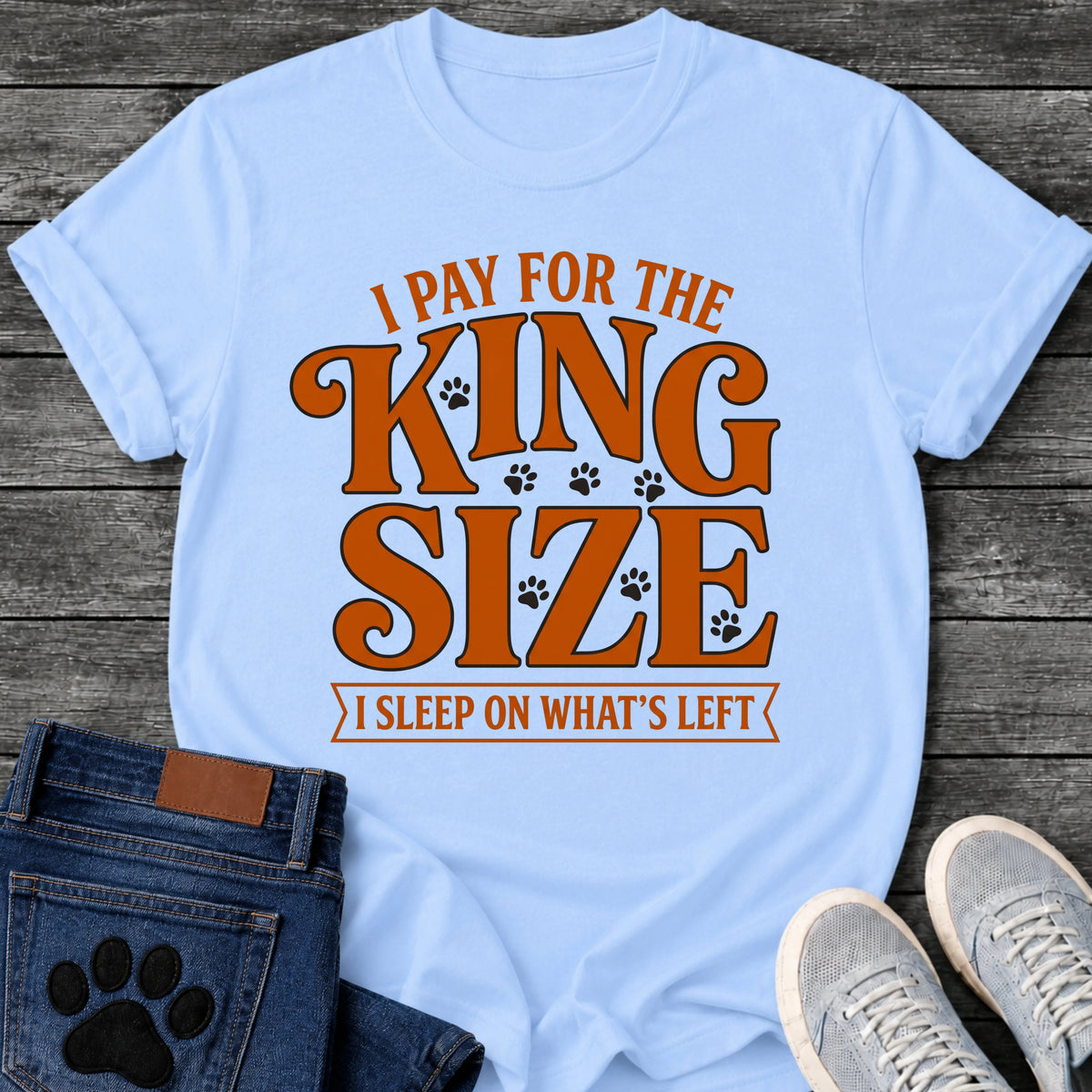 Bold vintage style graphic tee design reading "I Pay for the King Size, I Sleep on What's Left," in orange type with scattered paw prints and a banner underline.