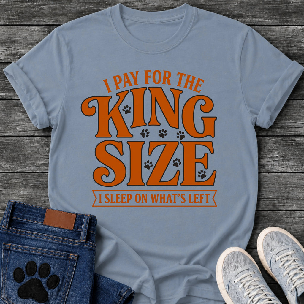 Bold vintage style graphic tee design reading "I Pay for the King Size, I Sleep on What's Left," in orange type with scattered paw prints and a banner underline.