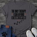 Bold graphic tee design reading "The Only Therapy I Can Afford Is Talking to My Dog," with orange accent lines and a trail of paw prints below.