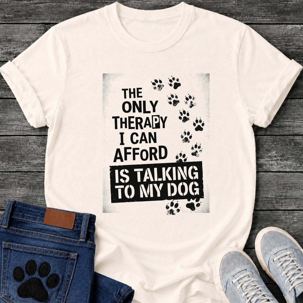 Bold stamped graphic tee design reading "The Only Therapy I Can Afford Is Talking to My Dog," with a trail of paw prints climbing the side.