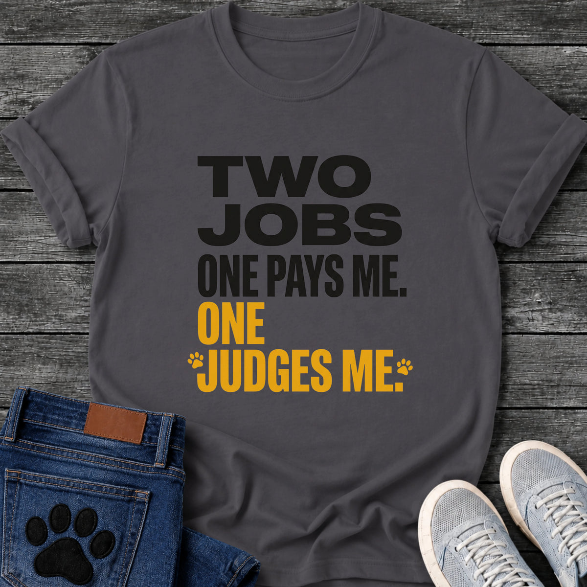 Bold two tone graphic tee design reading "Two Jobs, One Pays Me, One Judges Me," in black and orange with small paw prints framing the last line.