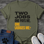 Bold two tone graphic tee design reading "Two Jobs, One Pays Me, One Judges Me," in black and orange with small paw prints framing the last line.