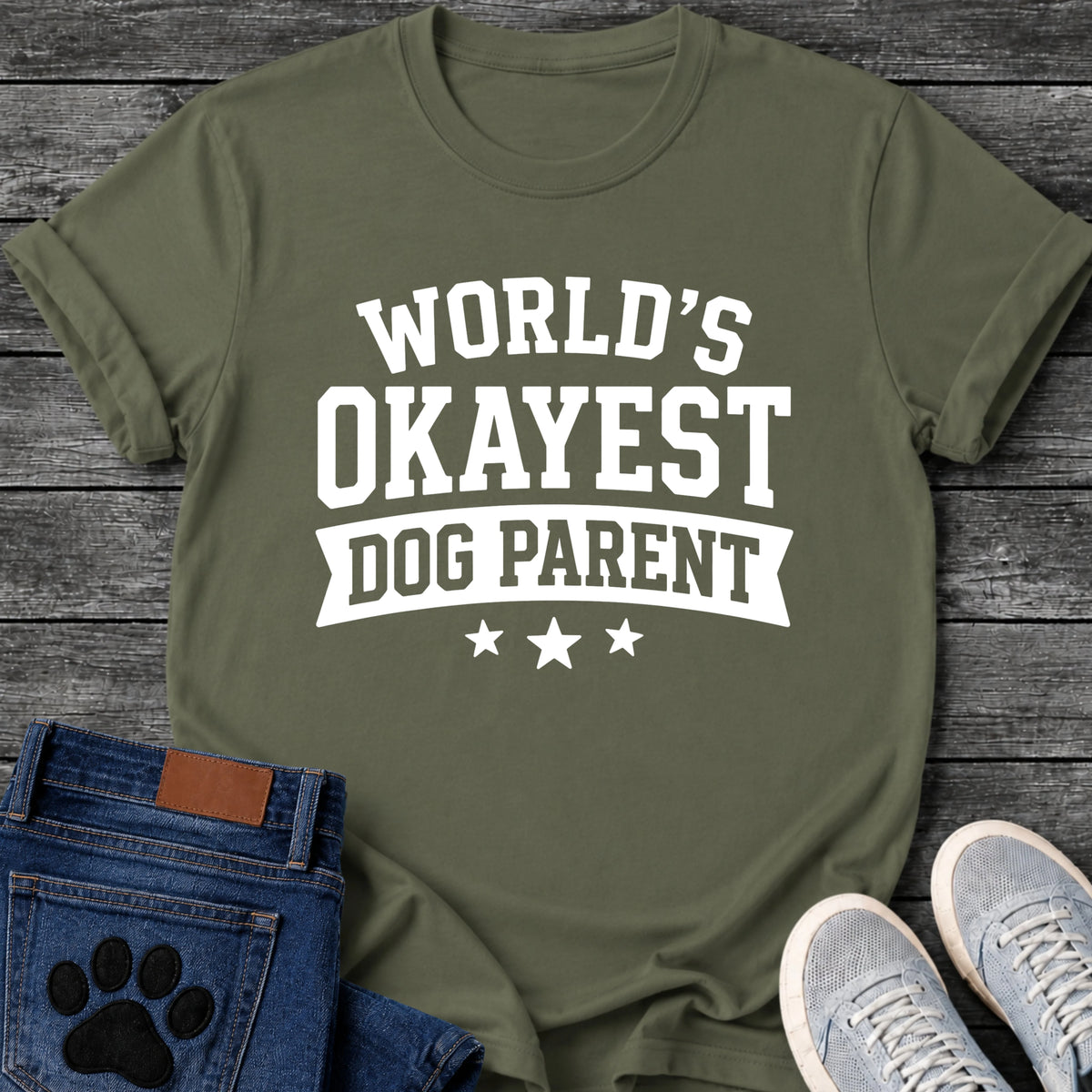 Varsity style graphic tee design in white lettering reading "World's Okayest Dog Parent," with a banner ribbon and three stars underneath.