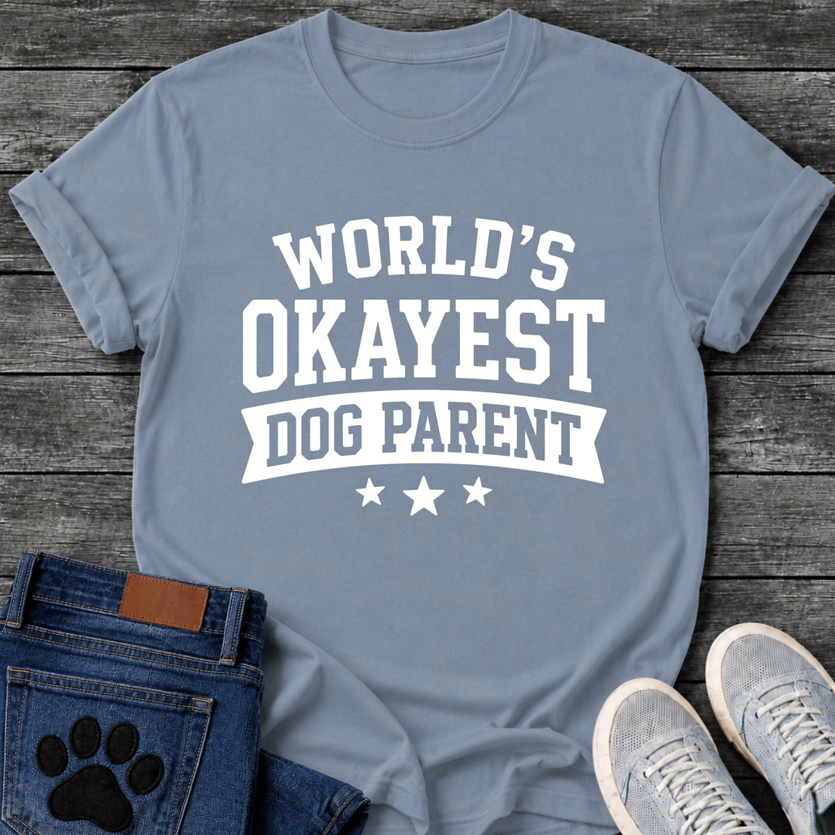 Varsity style graphic tee design in white lettering reading "World's Okayest Dog Parent," with a banner ribbon and three stars underneath.